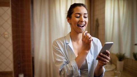 Frau hält beim Zähne putzen ihr Smartphone in der Hand