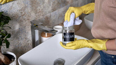 Frau steht vor Waschbecken und hält ein Glas mit VINEGAR-Beschriftung in der Hand und tunkt mit einem Tuch rein